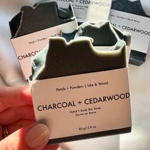 Load image into Gallery viewer, Hand holding a bar of soap labeled 'Charcoal + Cedarwood' with packaging in the background.