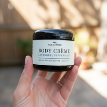 Load image into Gallery viewer, Hand holding a jar of Idle &amp; Wood Body Crème with a blurred outdoor background
