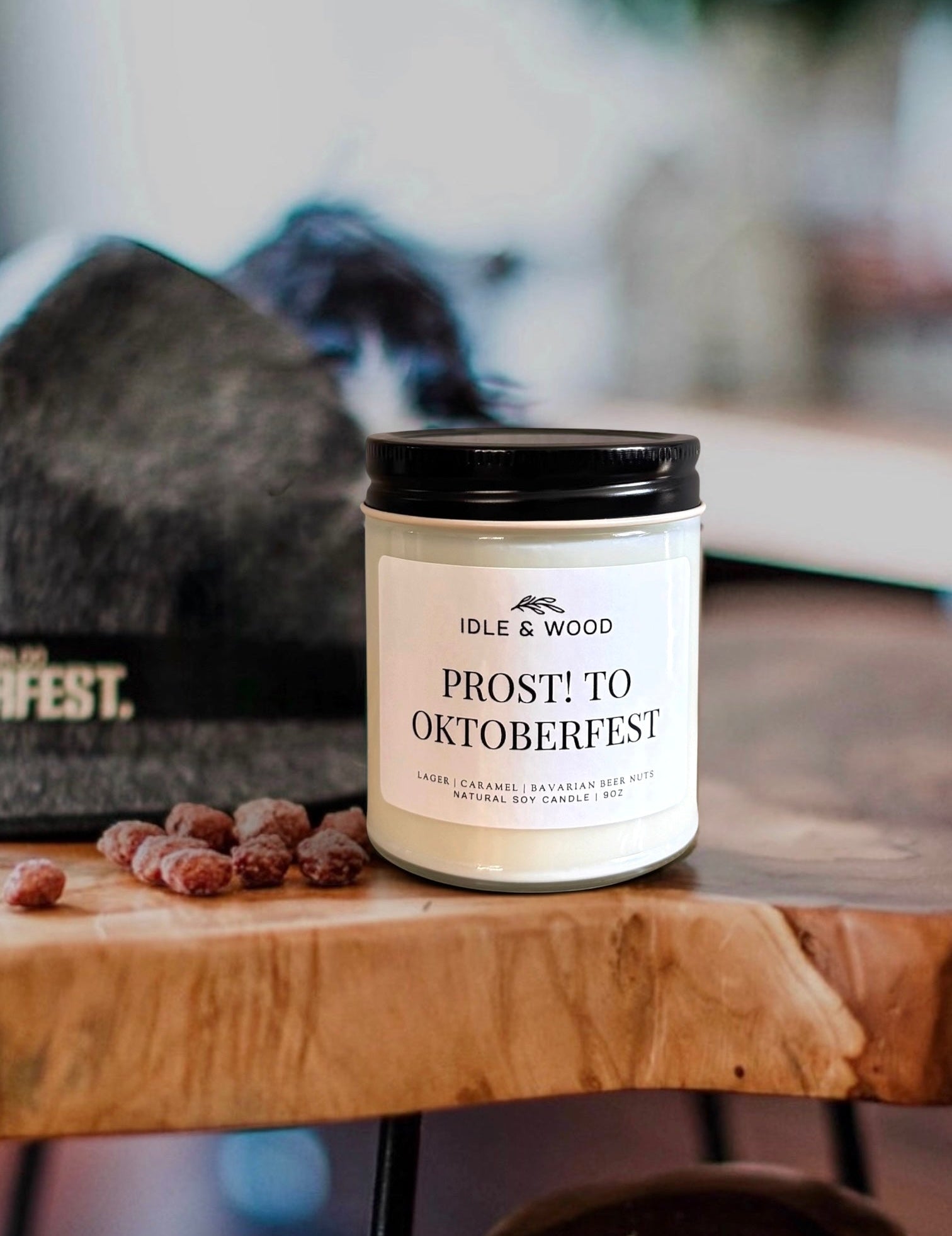A jar candle labeled 'PROST! TO OKTOBERFEST' by Idle & Wood sits on a wooden table next to an Oktoberfest hat with feather 