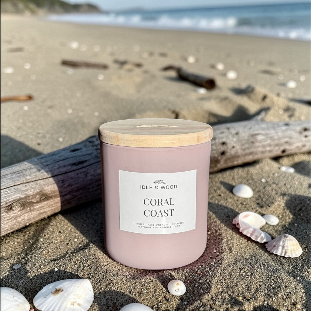 Candle labeled 'Coral Coast' by Idle & Wood on a sandy beach with shells and driftwood.